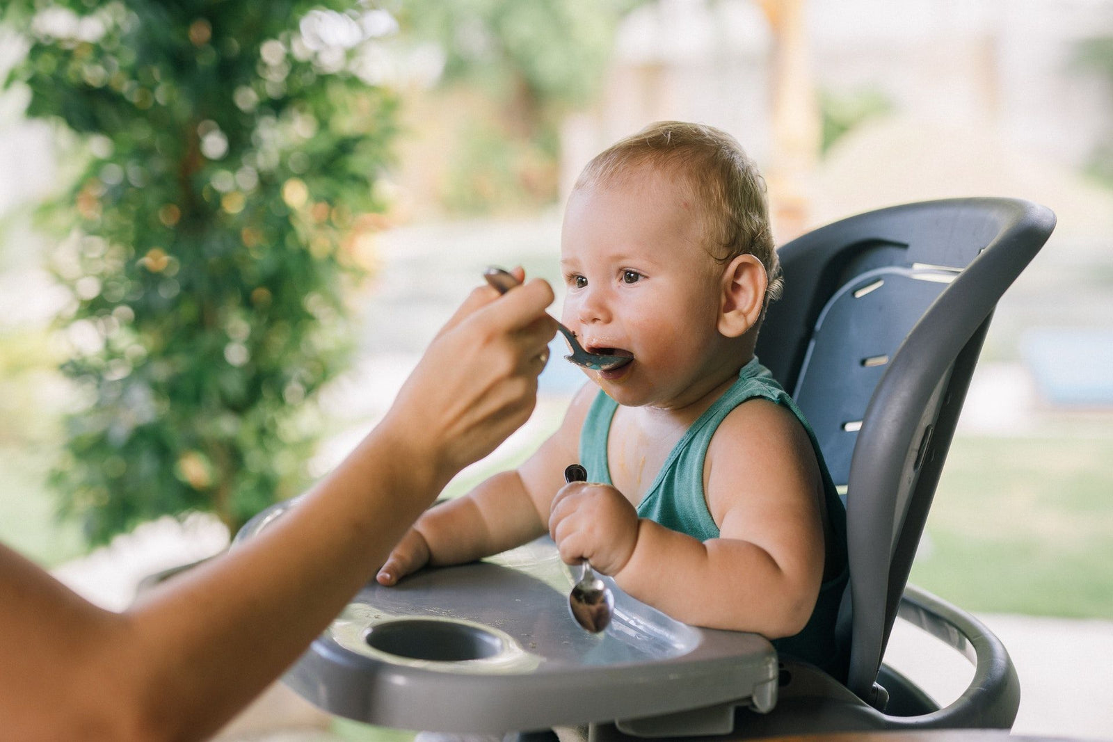 Ein Baby sitzt auf einem Kinderstuhl und bekommt von seiner Mutter Beikost zugeführt.