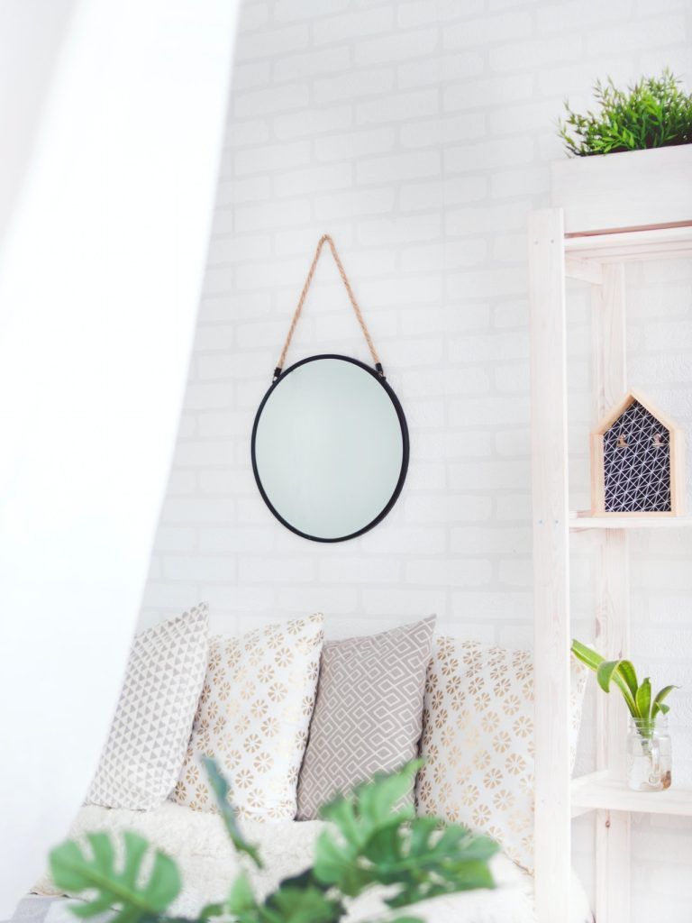 Ein Spiegel hängt an einer weißen Wand, darunter sind vier Kissen und eine grüne Pflanze davor: Alles dient zur gesunden Raumgestaltung.