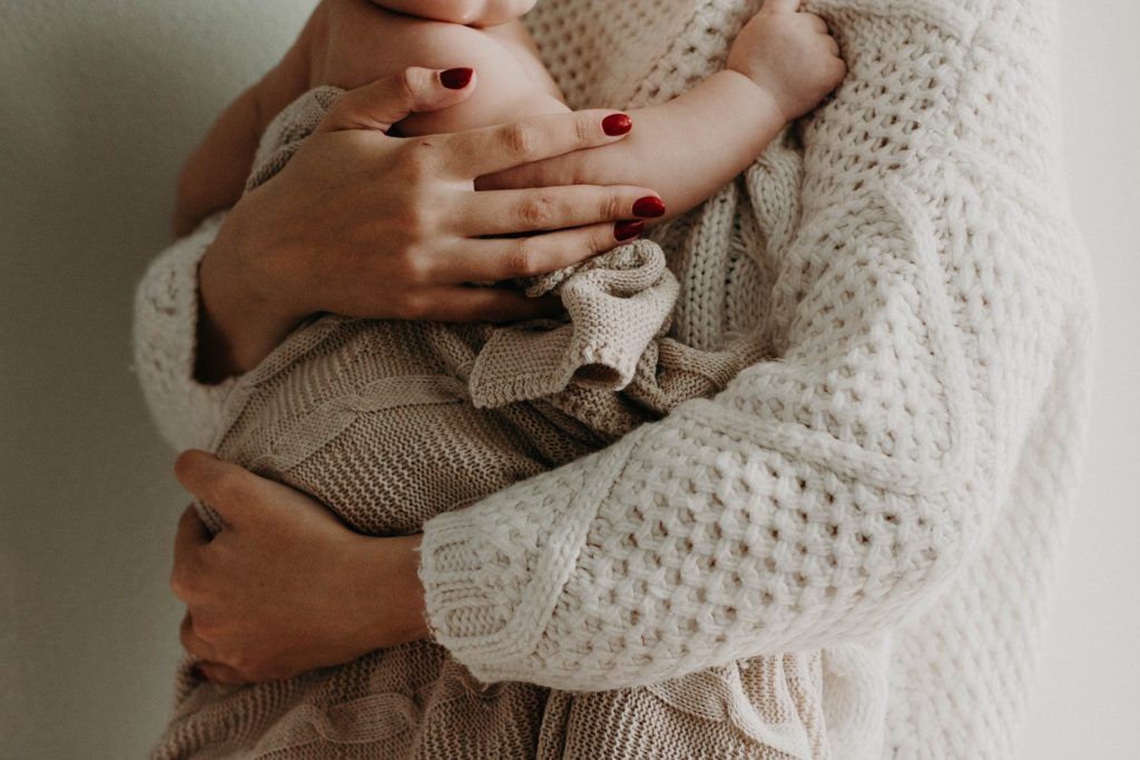 Eine Frau im weißen Pulli und roten Fingernägeln hält ein Baby, das Beikost bekommt, im Arm.